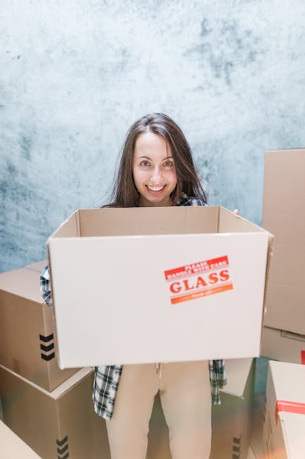 A smiling woman with curly black hair is sitting on a wooden floor surrounded by various cardboard moving boxes, some of which are open and partially filled with packing materials like white foam pean