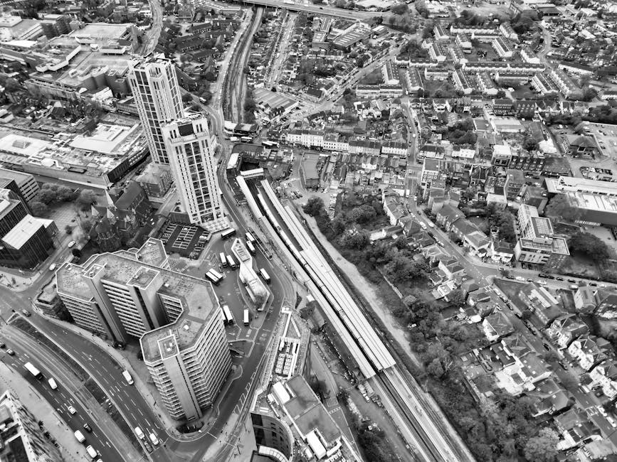 Aerial black and white photograph of a dense urban area with high-rise buildings, roads, and railway tracks. The image shows a cluster of multi-storey office and apartment buildings at the bottom left, with vehicles visible on the streets surrounding them. In the middle of the image, railway tracks run diagonally across, with an elevated train station platform visible along the tracks. The elevated platform is flanked by trees and residential houses on the right, with narrow streets and varied housing styles. Roads are busy with cars and buses, and the layout reveals a mix of commercial and residential structures. This scene illustrates a typical cityscape, emphasizing transportation infrastructure, vertical development, and urban density. Man with Van Croydon, providing removals and home relocation services, operates in such environments, managing furniture transport and packing logistics for moves near East Croydon station, Croydon.